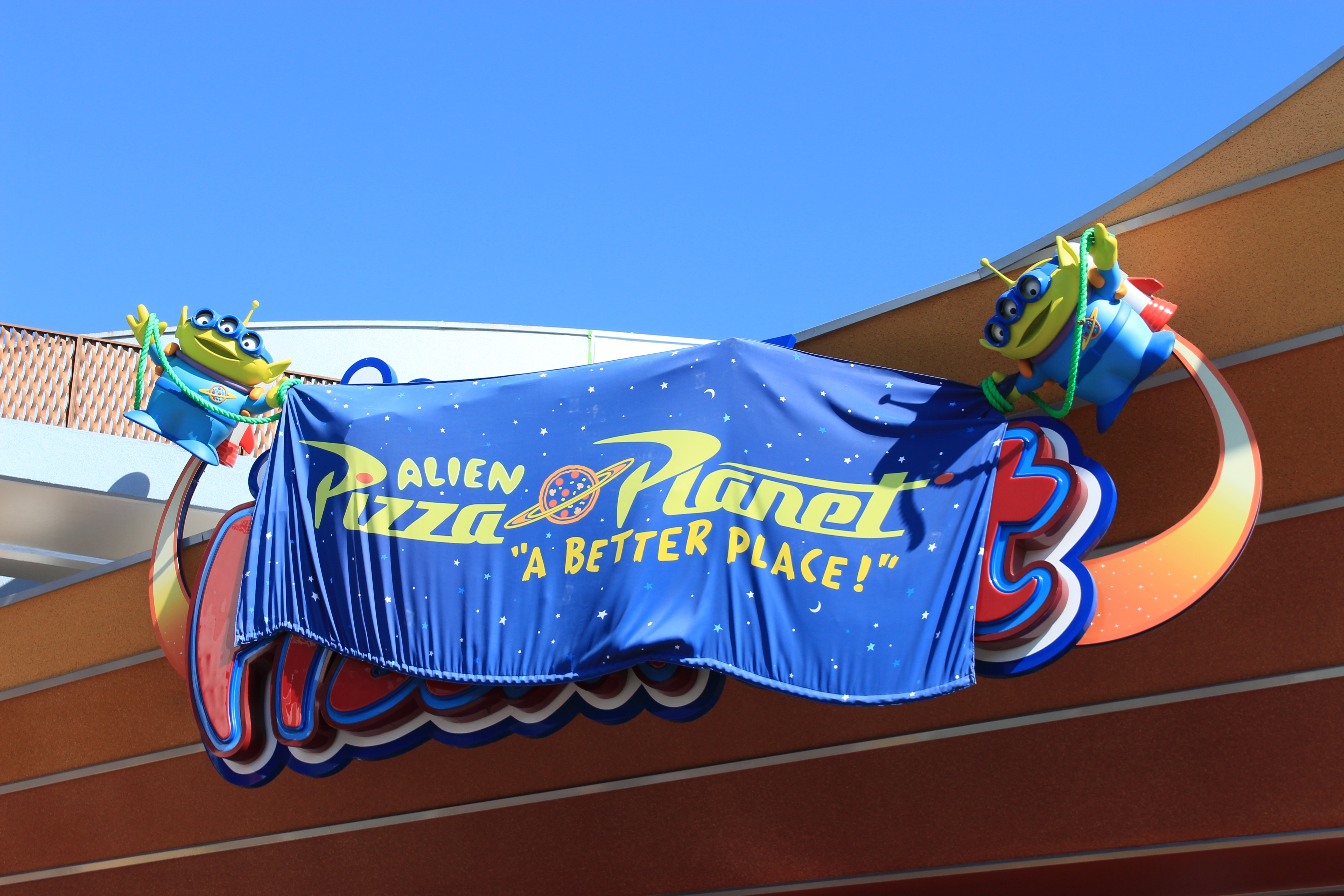 The sign for Alien Pizza Planet in Tomorrowland at Disneyland. It says "Alien Pizza Planet, A Better Place" on a cloth with two green aliens from Toy Story holding the cloth up.